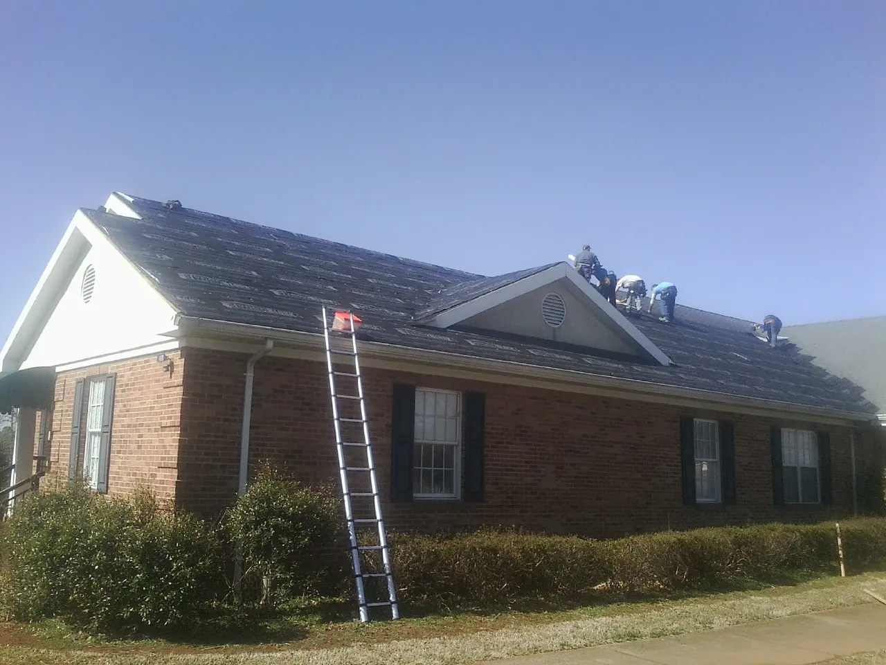 Crew tearing shingles off a brick ranch roof with a ladder set — Emergency Roof Repair work by our crew in Stapleton
