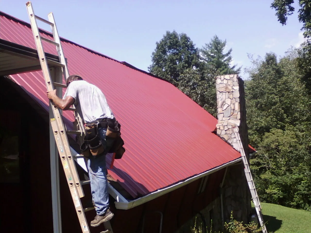 Roofer on a ladder beside a red metal roof and stone chimney — Soffit & Fascia Repair by Roofing Brothers in Stapleton